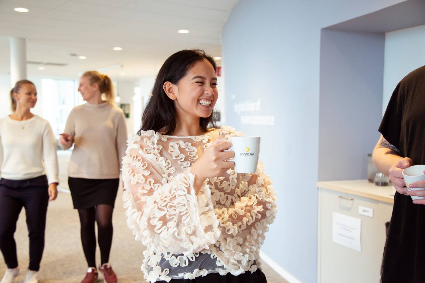 Smiling female colleague holding a coffee mug, sharing a laugh with teammates in the office